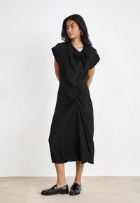 Robe noire, longueur genou, avec des manches courtes drapées ; détail de nœud torsadé à la taille ; tissu lisse et léger ; portée avec des mocassins noirs.