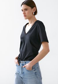 Ung kvinde med kort brunt hår iført sølv hoops øreringe, sort V-hals t-shirt og lyseblå jeans, stående med hænderne i lommen.
