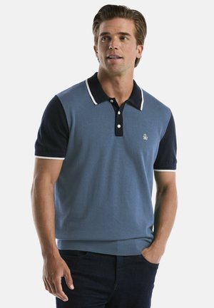 Mann mit braunen Haaren, der ein blaues Poloshirt mit dunkelblauen Ärmeln und Kragen trägt und mit einer Hand in der Tasche steht.