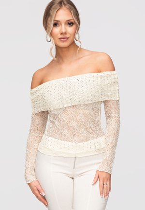 Femme aux cheveux blonds porte un top blanc en dentelle, manches longues et épaules dégagées, orné de perles, et un pantalon blanc sur un fond uni.