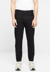 Pantalones de mezclilla negros con un corte recto, cinco bolsillos y cierre de botón. Llevados con zapatillas de colores claros.