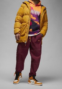 Giubbotto imbottito giallo, felpa con cappuccio grafica viola e arancione, pantaloni cargo color bordeaux e sneakers arancioni e nere con lo swoosh bianco.