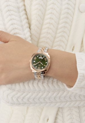 Zelta un sudraba toņu rokas pulkstenis ar zaļu ciparnīcu, romiešu skaitļiem un dimanta līdzīgiem akcentiem. Nēsāts uz rokas pret krēmkrāsas adīto džemperi.