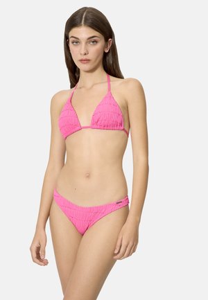 Joven mujer de pie con bikini rosa texturizado, top de triángulo y braguitas de talle bajo, sobre un fondo claro y sencillo.