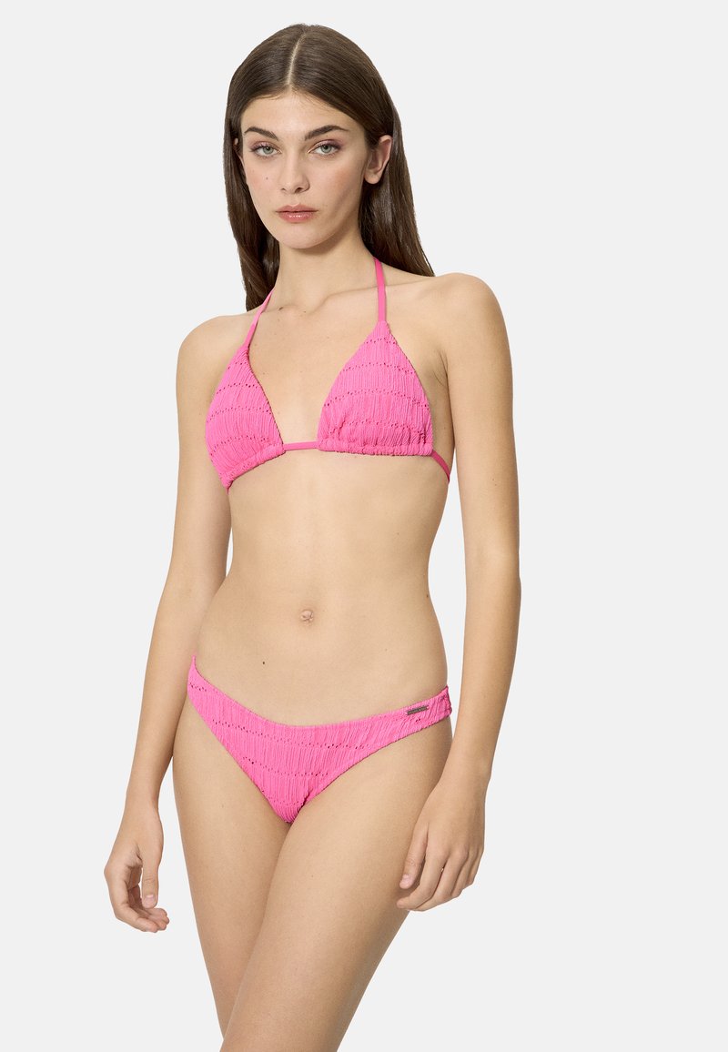 Joven mujer de pie con bikini rosa texturizado, top de triángulo y braguitas de talle bajo, sobre un fondo claro y sencillo.