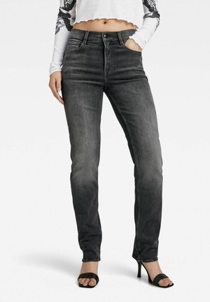 Frau trägt ausgewaschene schwarze High-Waist-Skinny-Jeans und schwarze Sandalen mit Absatz, dazu ein kurzes Langarmoberteil, das den Bauch freilegt.