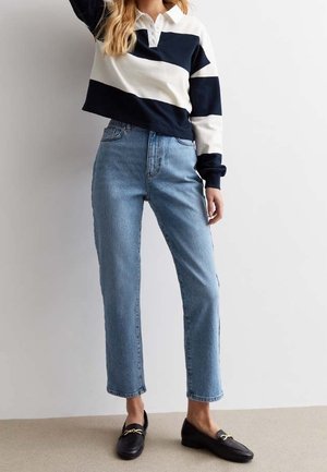 Vrouw met lichtblauwe rechtvallende spijkerbroek, zwarte loafers met gouden details, en een wit met marineblauw gestreept overhemd met lange mouwen en kraag.