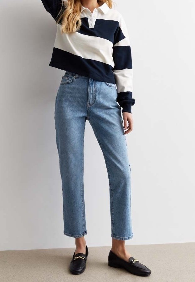 Femme portant un jean bleu clair droit, des mocassins noirs avec des détails dorés et une chemise blanche rayée bleu marine à manches longues et col.