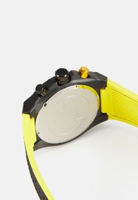 Reloj negro con caja redonda, correa de silicona amarilla y trasera de acero inoxidable. Presenta botones de presión negros y dorados a los lados.