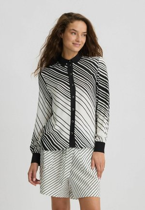 Blusa a maniche lunghe bianca e nera con strisce diagonali, colletto e bottone frontale. Abbinata a pantaloni corto a fantasia coordinati.
