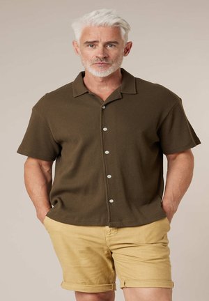 Homme aux cheveux et à la barbe blancs portant une chemise à manches courtes marron boutonnée et un short beige retroussé, debout les mains dans les poches.