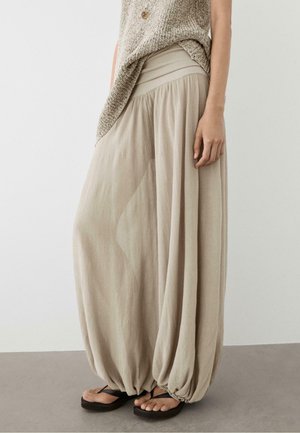 Woman wearing loose, beige harem pants with gathered ankles, paired with black flip-flops and a textured beige sleeveless top.
