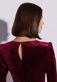 Bordowa sukienka z aksamitu z odkrytymi plecami w kształcie łezki, bufkowatymi rękawami i gładką fakturą. Projekt ma dopasowaną sylwetkę z dekoracyjnym skręceniem.