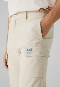 Beige corduroy cargo broek met een zijzak, voorzien van een blauw Red Bull Racing logo patch en een gestructureerd rib materiaal.