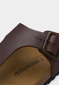 Καφέ λουρί δερμάτινου σανδαλιού με υφή, με φθαρμένη ορείχαλκο πόρπη πάνω σε ταμπά πάτο Birkenstock και μαύρη σόλα.