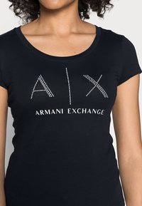 Černé bavlněné tričko s kulatým výstřihem, zdobené bílým logem "A|X ARMANI EXCHANGE" s kamínky. Vytvarovaný střih se short rukávy.