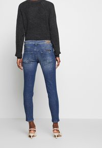 Suéter gris oscuro y jeans ajustados azules, con un bolsillo trasero con detalle de cremallera y una parche de cuero en la cinturilla. Zapatos en tonos neutros.