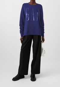Sudadera morada con "GAP" en letras reflectantes, combinada con pantalones anchos de rayas negras y botas negras de tobillo; sosteniendo un bolso blanco.