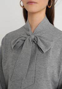 Pull en tricot gris avec un col montant et un grand nœud noué au niveau du cou. Texture douce avec des lignes lisses et épurées.