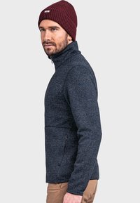 Marineblaue Fleecejacke mit hohem Kragen, strukturiertem Muster, Brusttasche und seitlichen Reißverschlüssen. Kombiniert mit einer burgunderfarbenen gerippten Mütze.