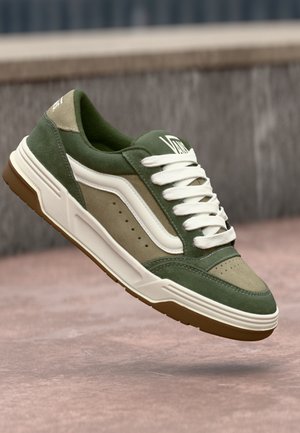 Sneaker verde e beige con texture in suede e pelle, lacci bianchi e suola in gomma. Presenta perforazioni sui lati e un marchio sulla linguetta.