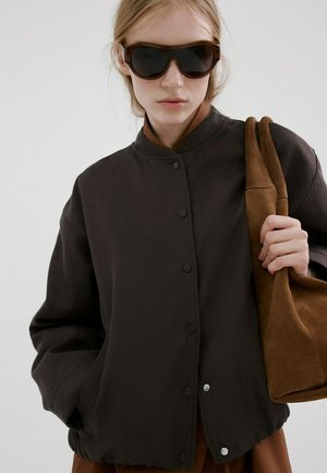 Jeune femme portant de grandes lunettes de soleil noires, veste noire boutonnée, tenant un grand sac en daim marron sur l'épaule.