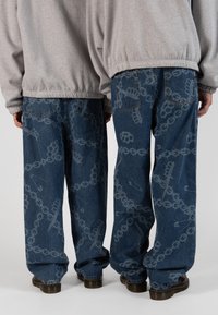 Jeans de mezclilla azul holgados con un diseño estampado que presenta cadenas y alfileres de seguridad, complementados con una sudadera gris y zapatos negros.