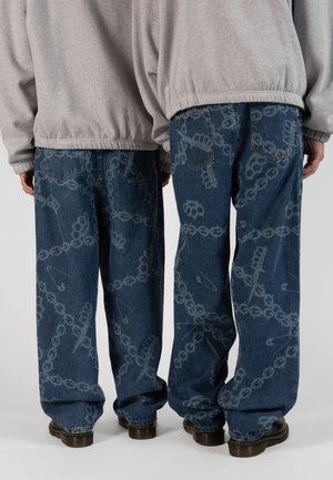 Baggy blå denim jeans med et mønster, der indeholder kæder og sikkerhedsnåle, suppleret af en grå sweatshirt og sorte sko.