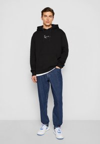 Svart hettegenser med frontlomme og hvit logo, parret med blå denimjeans og hvite sneakers med blå og svarte detaljer.