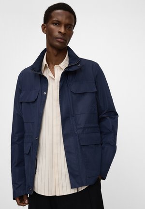 Mann trägt eine marineblaue Arbeitsjacke über einem beige-vertikal gestreiften Hemd und steht vor einem schlichten weißen Hintergrund.