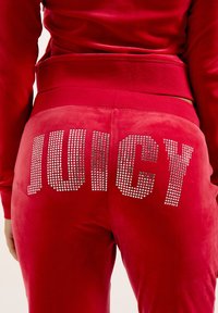 Rote Velour-Sweatpants mit großen Strassbuchstaben, die "JUICY" auf der Rückseite buchstabieren, mit einer glatten, weichen Textur und einem elastischen Taillenbund.