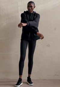 Fekete kapucnis pulóver elülső cipzárral, fekete leggings-szel és fekete sportcipővel, fehér talppal kombinálva. Az anyag simának és testhezállónak tűnik.