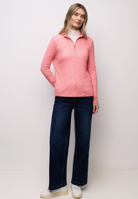 Vaaleanpunainen vetoketjullinen neulepaita pitkähihainen, ribbineulosta, yhdistetty leveälahkeisiin tummansinisiin farkkuihin ja valkoisiin lenkkareihin. Ei näkyviä kuvioita tai yksityiskohtia.