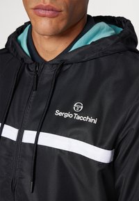 Chaqueta negra con capucha que cuenta con una franja horizontal blanca, tejido texturizado y un logotipo. El interior de la capucha es de color turquesa, añadiendo un contraste de color.