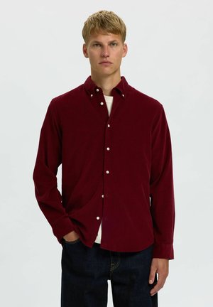 Jeune homme aux cheveux blonds courts portant une chemise bordeaux boutonnée sur un t-shirt blanc et un jean foncé, debout devant un fond uni clair.
