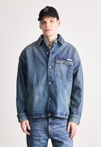 Τζάκετ τζιν σε ανοιχτό μπλέ με γιακά, μπροστινό κλείσιμο με κουμπιά και τσέπη με πατάκι. Φορεμένο με τζιν σε ταιριαστό μπλέ και μαύρο καπέλο.