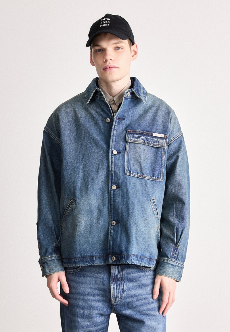 Τζάκετ τζιν σε ανοιχτό μπλέ με γιακά, μπροστινό κλείσιμο με κουμπιά �και τσέπη με πατάκι. Φορεμένο με τζιν σε ταιριαστό μπλέ και μαύρο καπέλο.