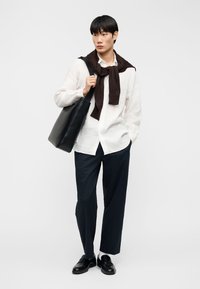 Jeune homme portant une chemise blanche à boutons, un pantalon noir, des chaussures noires, un pull marron posé sur les épaules, portant un grand sac cabas noir à l'épaule.