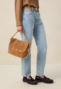 Sac à main en daim marron avec un fermoir à rabat et trois clous décoratifs en métal, associé à un jean droit bleu clair et des mocassins foncés.