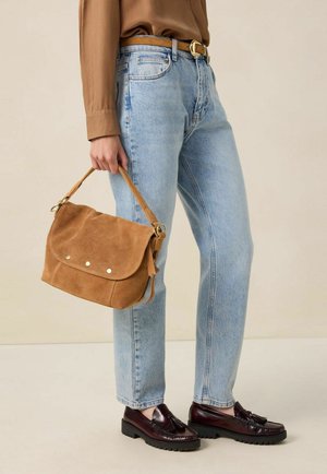 Sac à main en daim marron avec un fermoir à rabat et trois clous décoratifs en métal, associé à un jean droit bleu clair et des mocassins foncés.