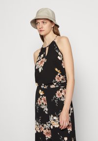 Vestido preto floral com decote halter, apresentando um detalhe em forma de chave, e acentuado com padrões de flores bege. Usado com um chapéu de balde bege.