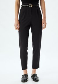 Hoge zwarte broek met een op maat gemaakte pasvorm, voorzien van voorplooien en een zwart leren riem met een gouden gesp, gecombineerd met glanzende zwarte loafers.