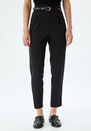 Pantalones negros de cintura alta con un corte ajustado, que presentan pliegues frontales y un cinturón negro con hebilla dorada, combinados con mocasines negros brillantes.