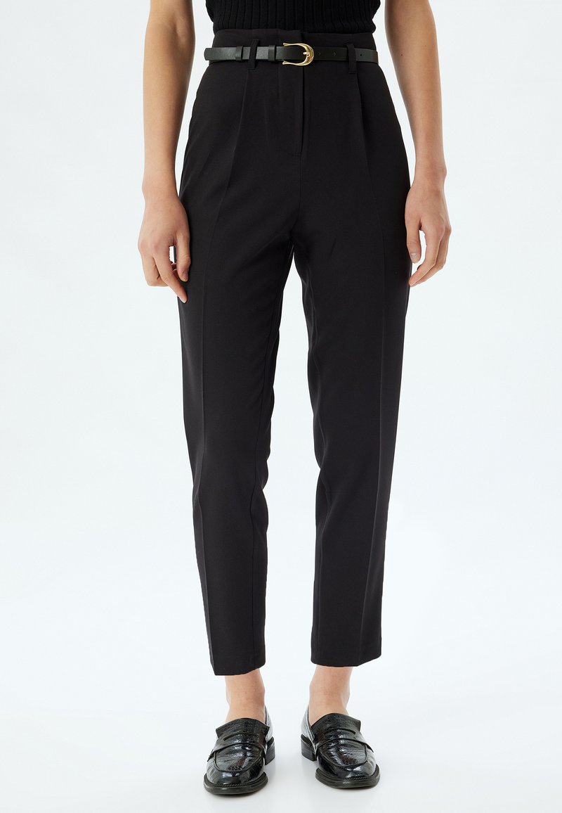 Hoge zwarte broek met een op maat gemaakte pasvorm, voorzien van voorplooien en een zwart leren riem met een gouden gesp, gecombineerd met glanzende zwarte loafers.