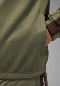 Olivgrön träningsjacka med en ficka, elastiska muddar och texturerat tyg. Inkluderar en brun detalj med vit text.