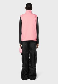 Rosa ärmlös väst över en svart långärmad tröja; kombinerat med vida svarta byxor med cargo-fickor, samt en liten rosa accessoar.