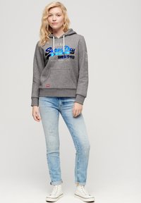 Sudadera gris con bolsillo frontal, cordones blancos y logo colorido de "Superdry". Pantalones vaqueros ajustados desgastados en azul claro y zapatillas blancas.