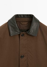Bruine jas met een zwarte leren kraag en knoopsluiting aan de voorkant. Het materiaal is glad met een matte textuur.