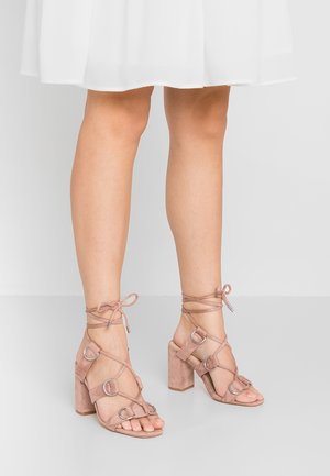 Blush pink suede block-heeled sandals with multiple straps, featuring circular hardware accents and ankle ties, set against a white background.