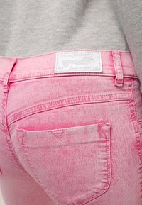 Lähikuva haalistuneista vaaleanpunaisista farkuista takataskussa, jonka yllä on valkoinen Diesel Industry -labeli harmaan yläosan päällä.